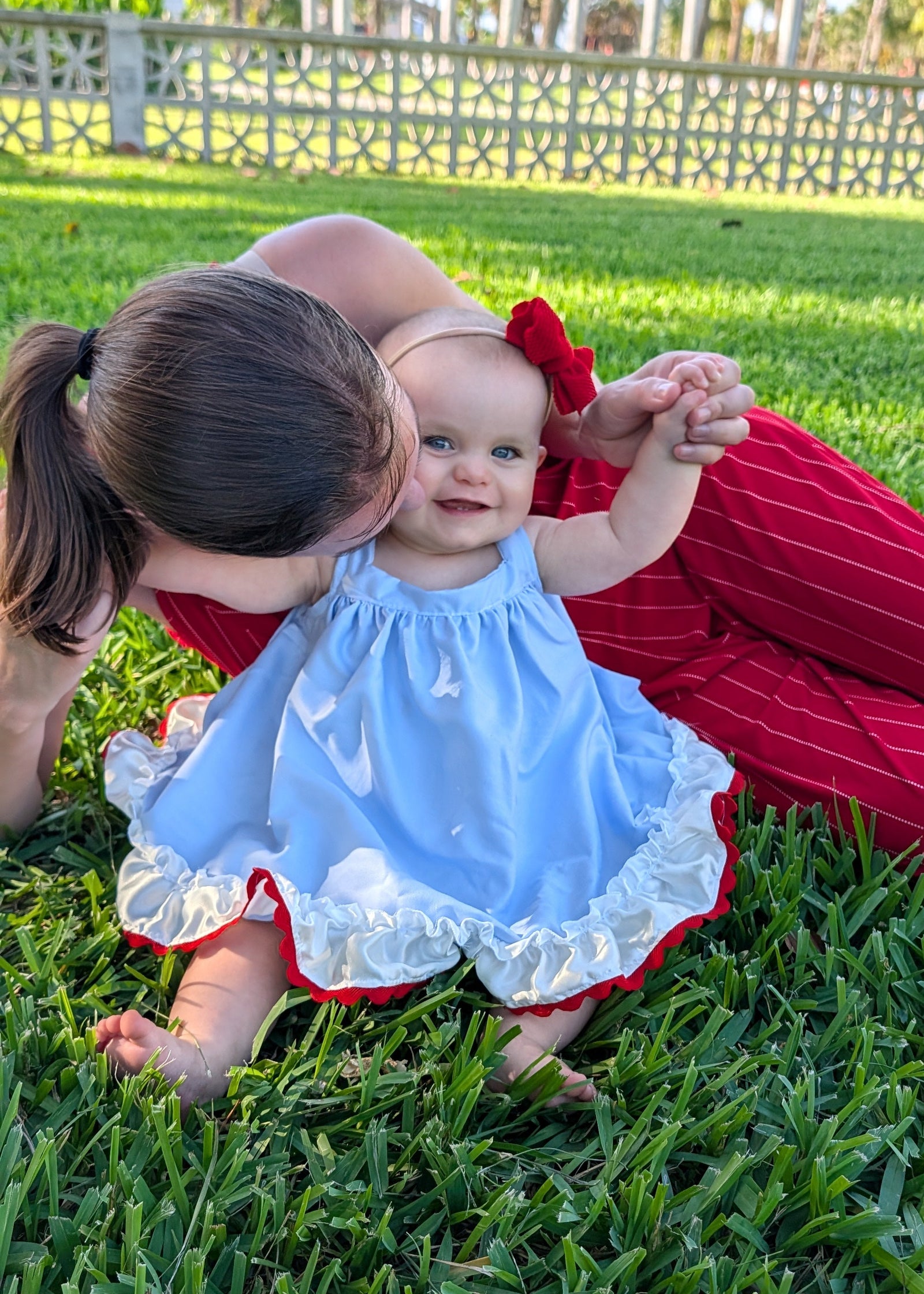 A baby girl wearing a light blue sleeveless dress with white ruffles and a red bow sits on green grass while a woman in red pinstriped pants leans in close to her.