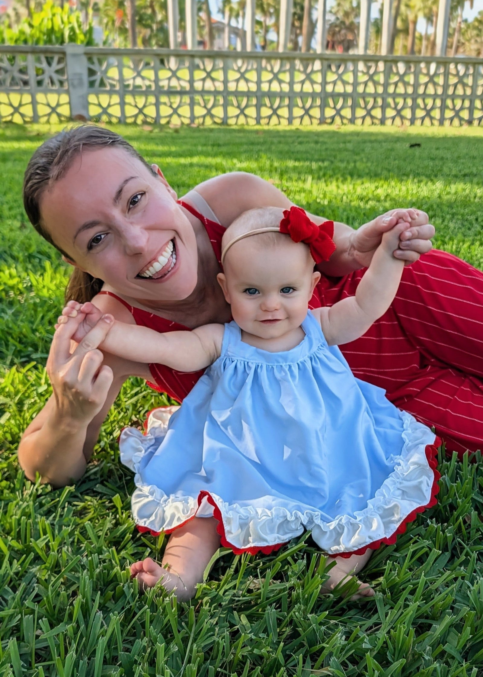 A smiling woman and a baby sitting together on green grass outdoors. The baby is wearing a light blue sleeveless dress with white ruffle trim and a red bow headband, while the woman wears a red outfit.