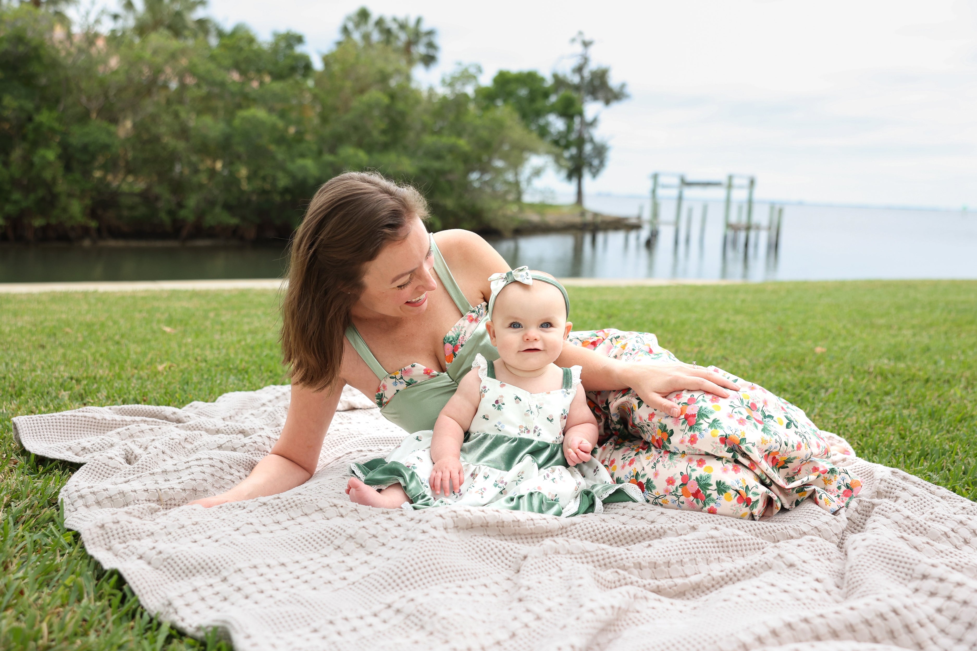 A woman and a baby lying on a white crocheted blanket on a grassy lawn, with water and trees in the background. The woman wears a green floral dress, and the baby wears a matching green and white floral dress.