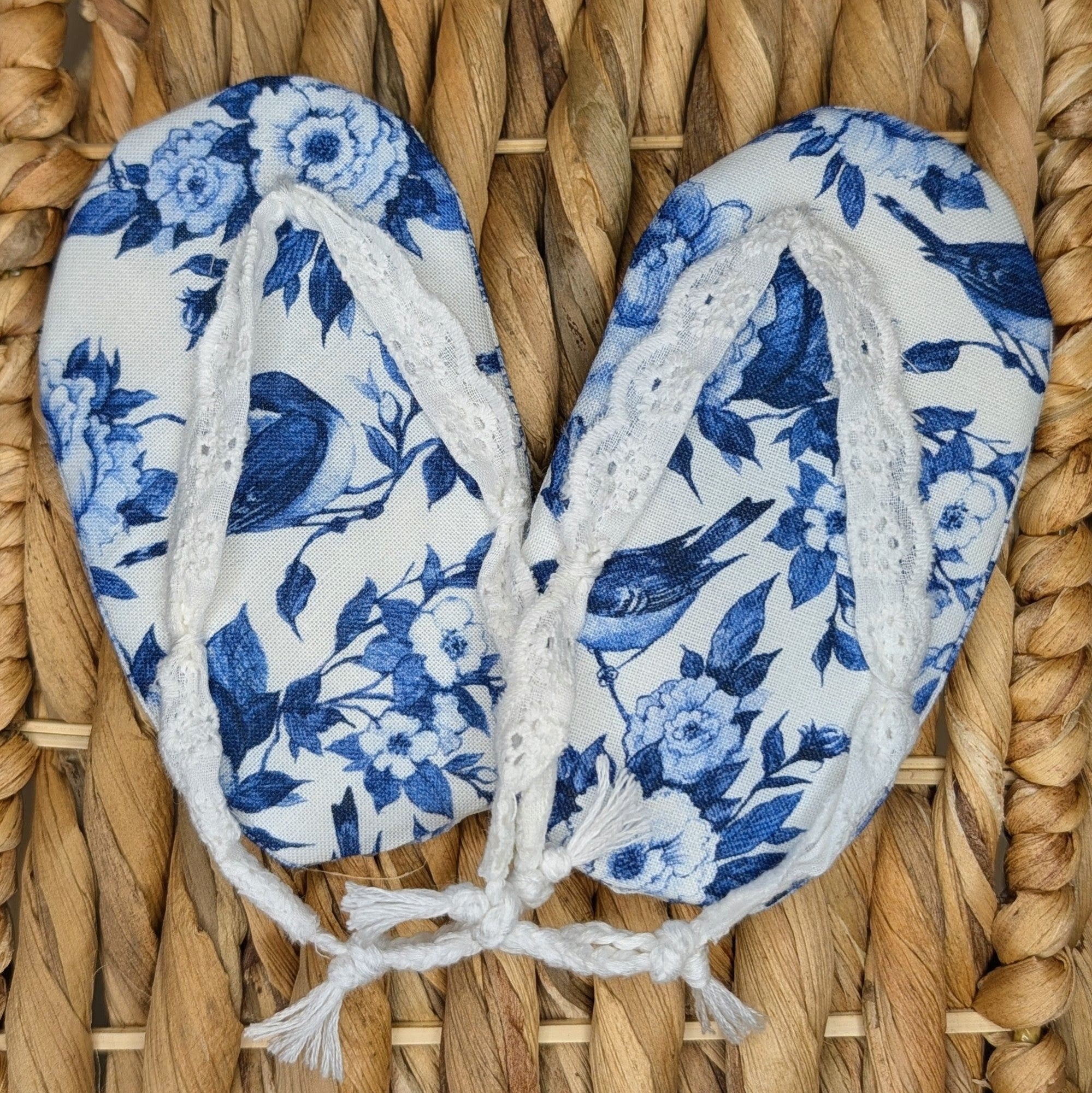 A pair of blue and white floral print thong sandals with white lace straps, laid flat on a woven straw mat.