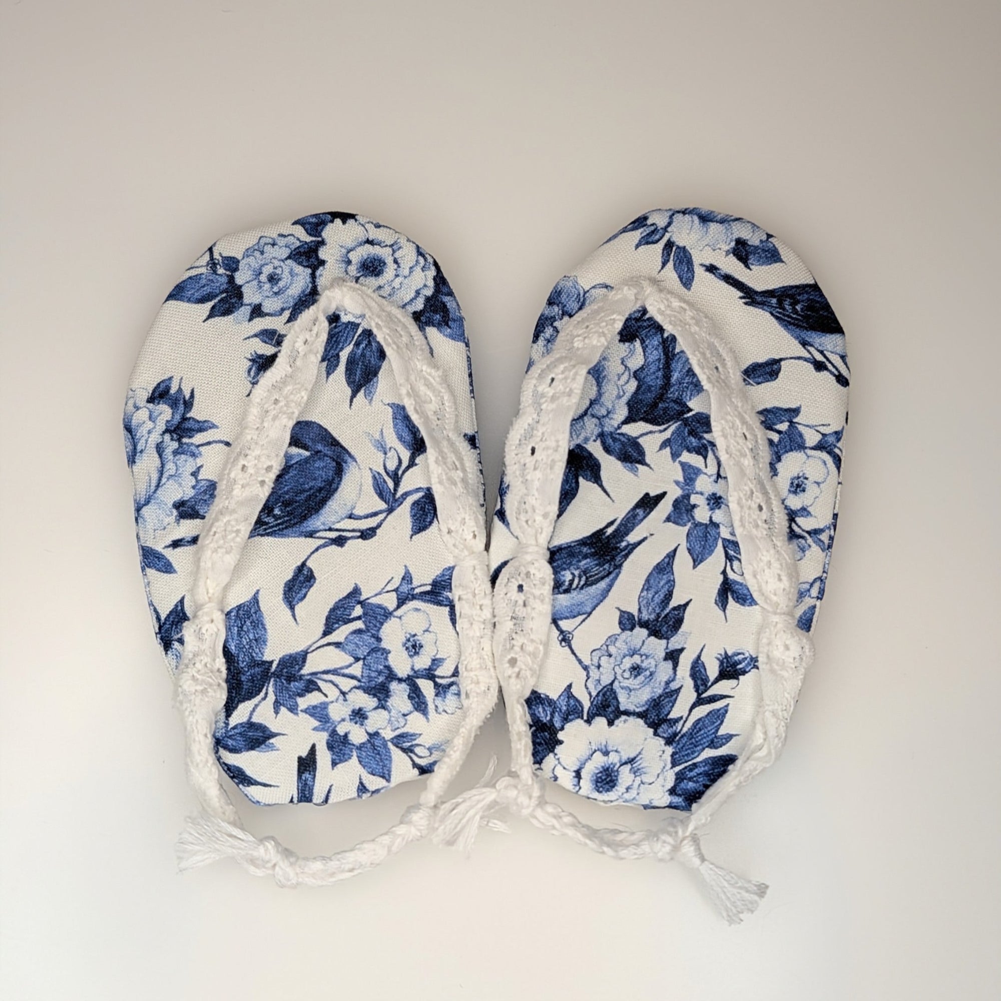 A pair of baby flip-flops with a blue and white floral print and white lace trim, laid flat on a light off-white background.