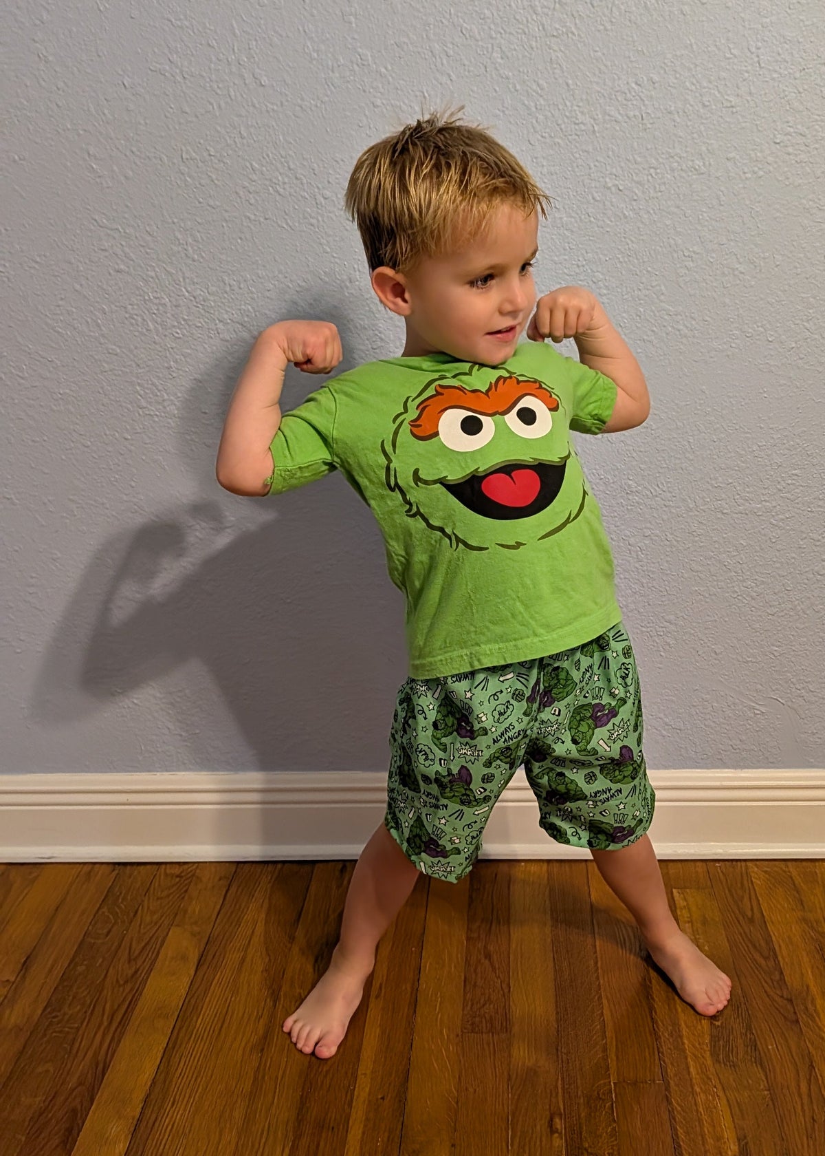 A young boy posing in a green Hulk shorts, standing on a wooden floor against a light gray wall.