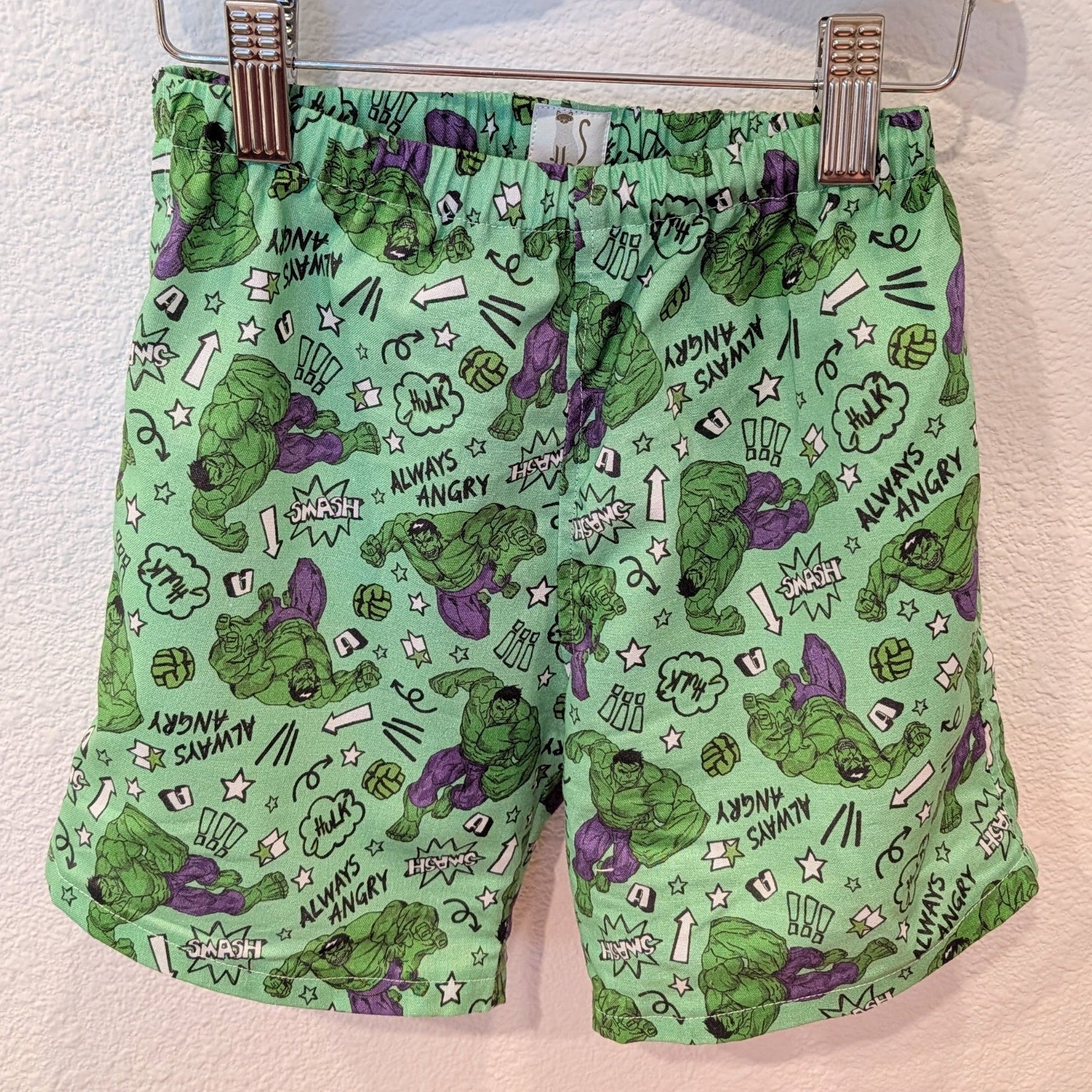 Green shorts featuring a comic-book style print of the Hulk, including angry expressions, 'ANGRY SAMMY' text, and comic-style doodles, hanging from a hanger with metal clips against a textured wall.