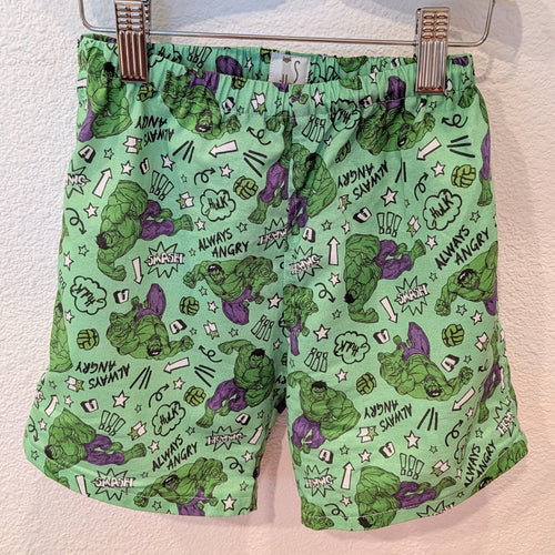Green shorts featuring a comic-book style print of the Hulk, including angry expressions, 'ANGRY SAMMY' text, and comic-style doodles, hanging from a hanger with metal clips against a textured wall.