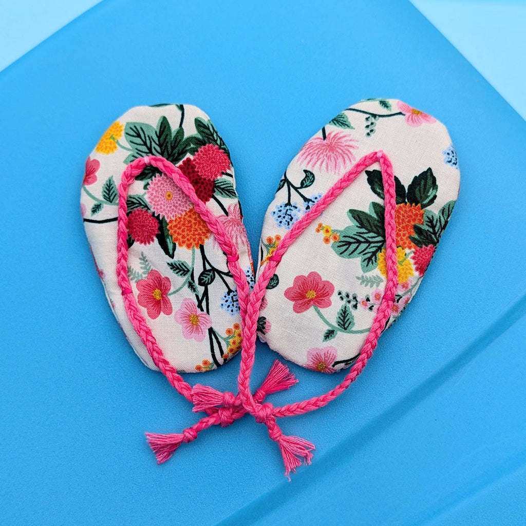 A pair of miniature floral flip-flop sandals with pink straps, arranged on a bright blue surface.