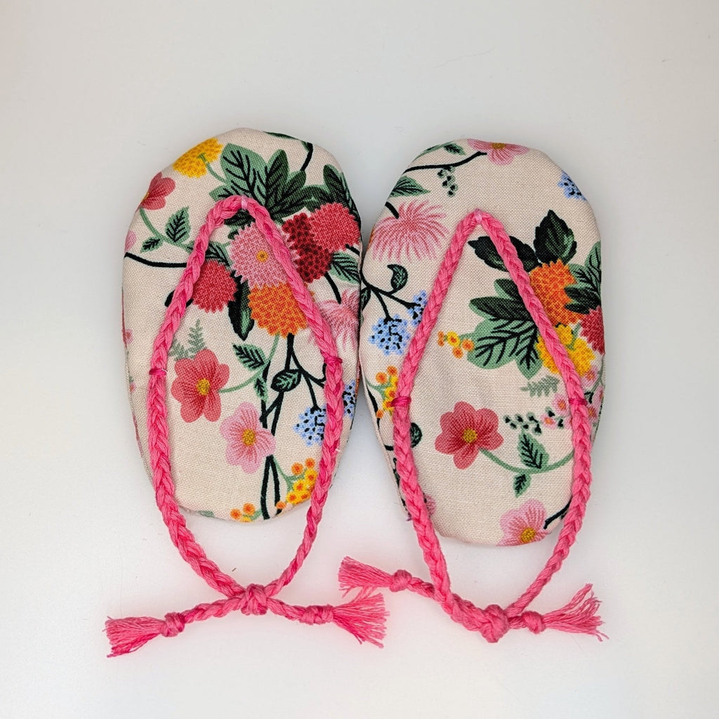 A pair of handmade flip-flop sandals with a colorful floral pattern on a cream background and bright pink braided straps with tassels, shown on a plain white background.