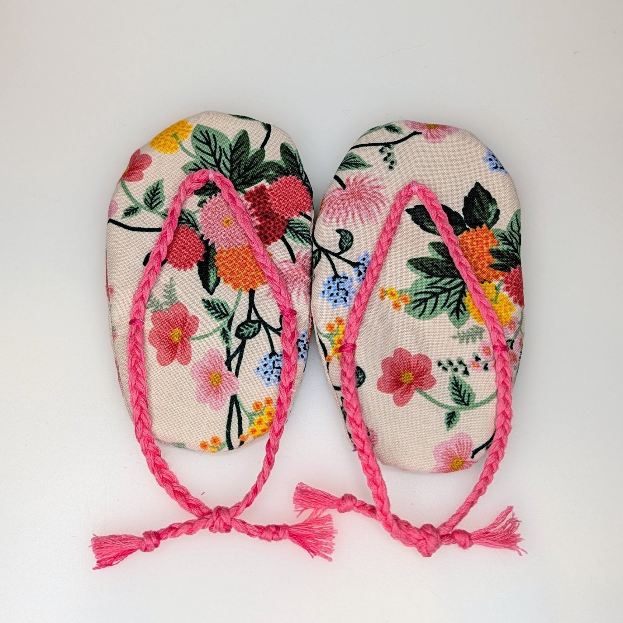 A pair of handmade flip-flop sandals with a colorful floral pattern on a cream background and bright pink braided straps with tassels, shown on a plain white background.