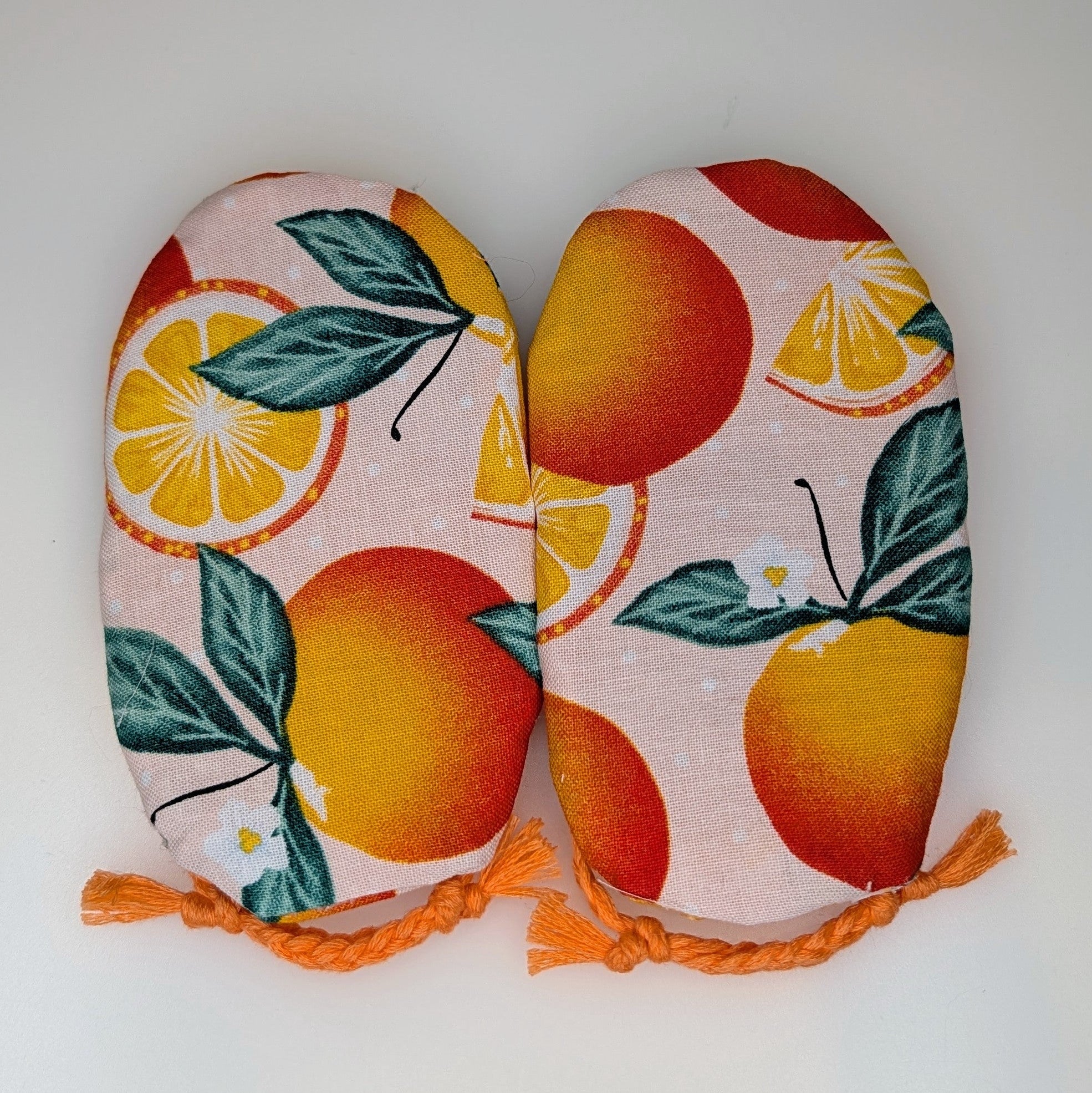 A pair of fabric oven mitts featuring a vibrant citrus print with oranges, lemons, and green leaves on a light pink background, finished with orange tassels.