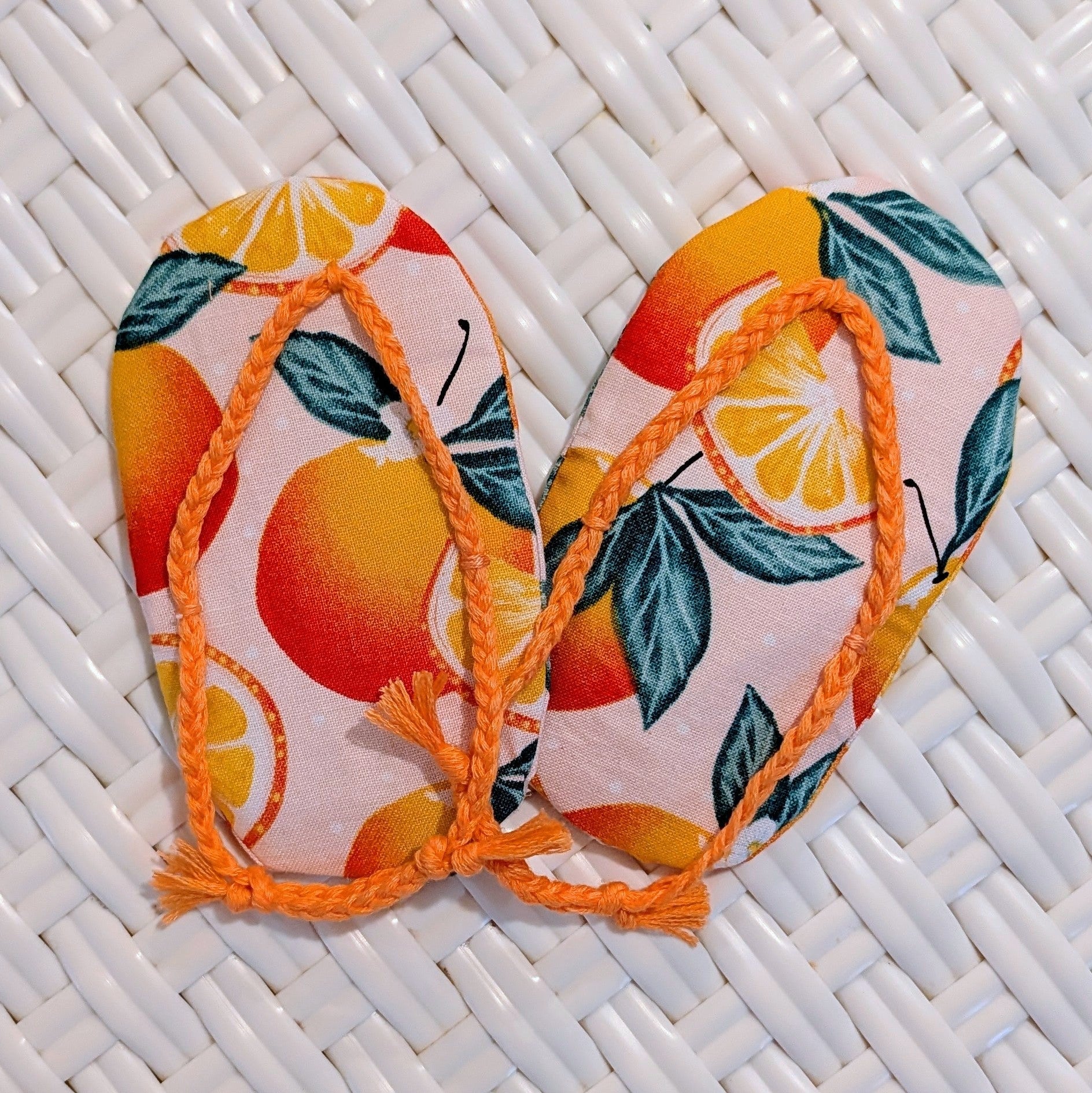 A pair of miniature flip-flop sandals with a citrus orange and green leaf pattern on a light pink background, resting on a white woven mat.