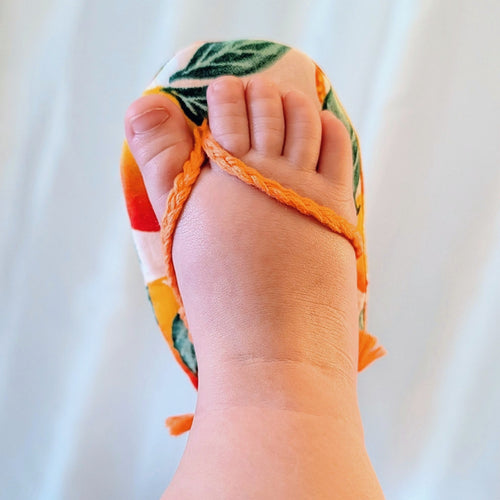 A close-up of a foot wearing a colorful, open-toe sandal with orange braided straps and a tropical leaf patterned upper, set against a soft white curtain background.