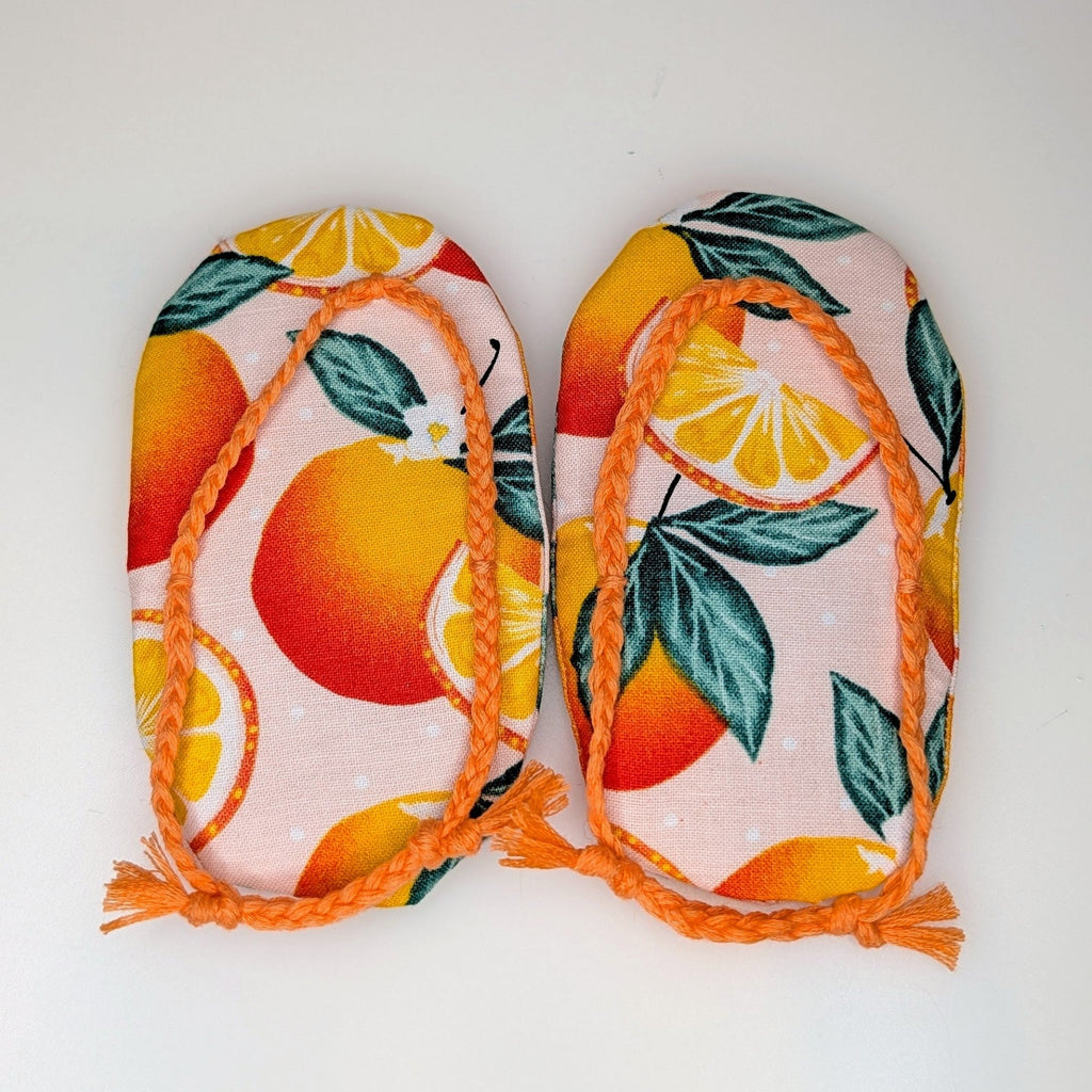 A pair of handmade fabric flip-flops featuring an orange and green citrus pattern on a light pink background, finished with orange braided straps and tassels, displayed on a plain white surface.