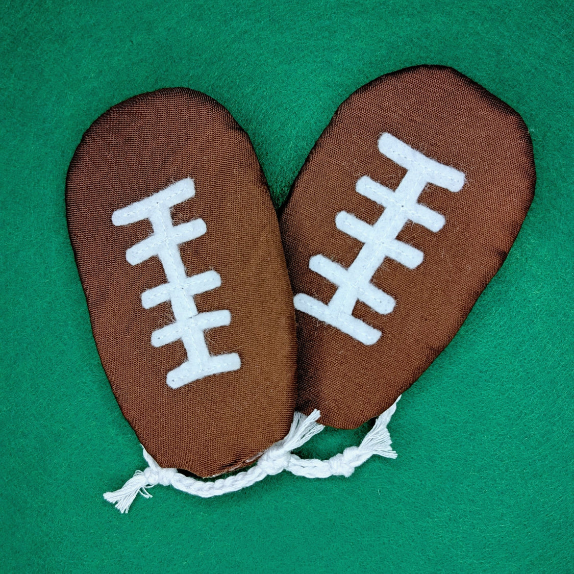 A pair of brown football-shaped flip-flops with white laces, laid flat on a green felt surface.
