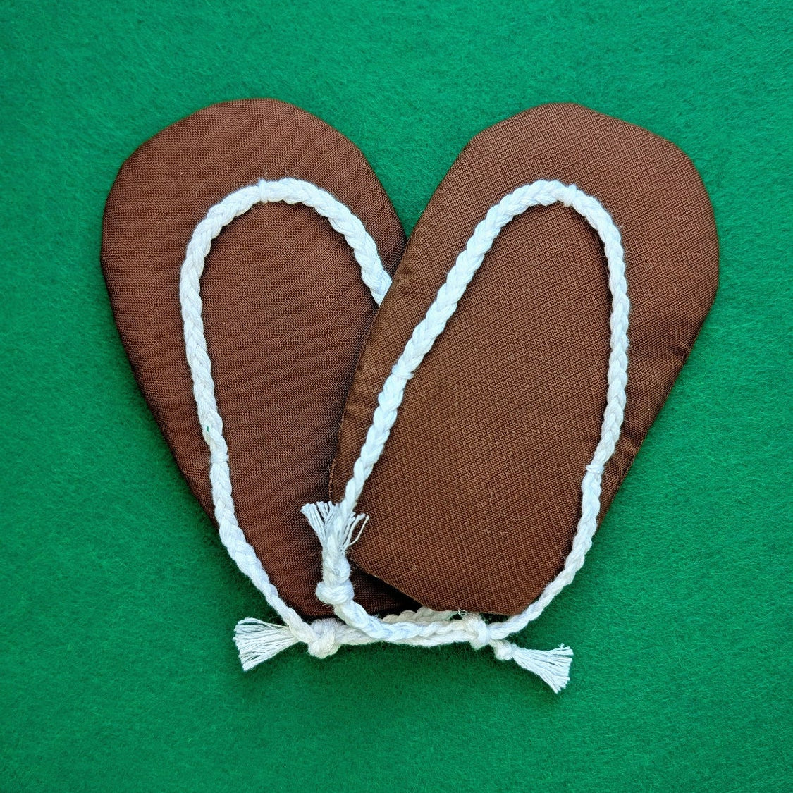 A pair of brown fabric slip-on flip-flops with white braided rope trim and tassels, laid flat on a solid green background.