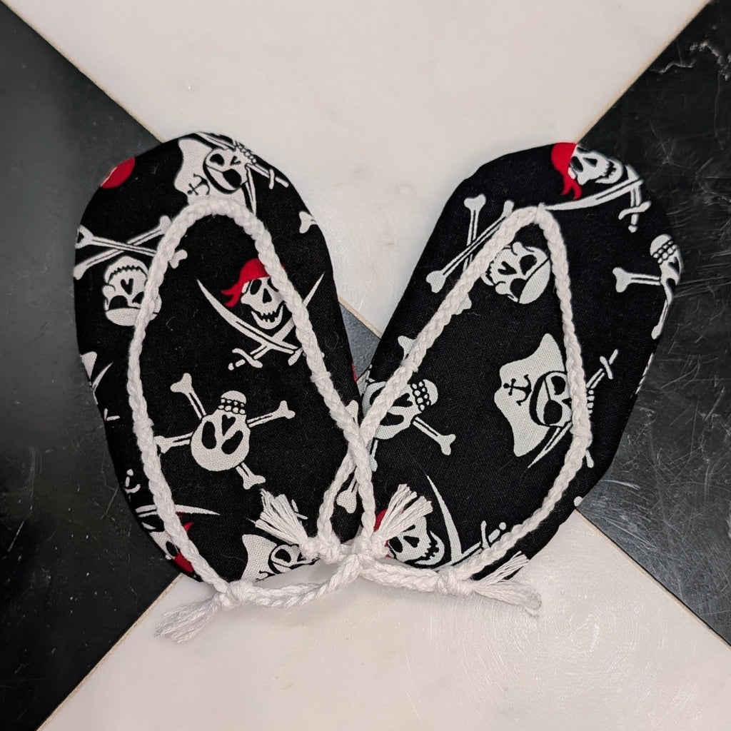 Black pirate-themed fabric accessories with white skull and crossbones print and white rope trim, tied together at the center on a black and white marble surface.
