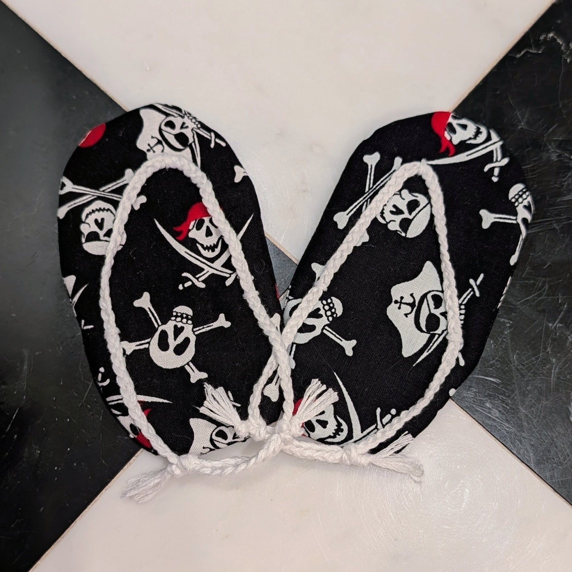 Black pirate-themed fabric accessories with white skull and crossbones print and white rope trim, tied together at the center on a black and white marble surface.