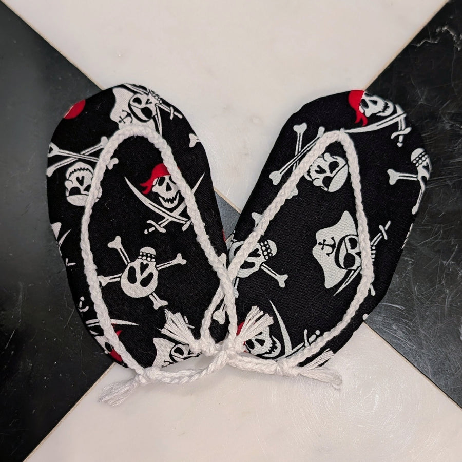Black pirate-themed fabric accessories with white skull and crossbones print and white rope trim, tied together at the center on a black and white marble surface.