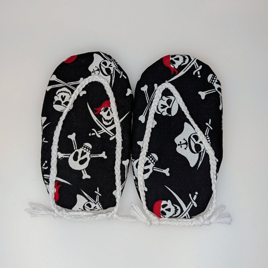 A pair of black flip-flop slippers with a white skull and crossbones pirate print and white fringe trim, laid flat on a white background.
