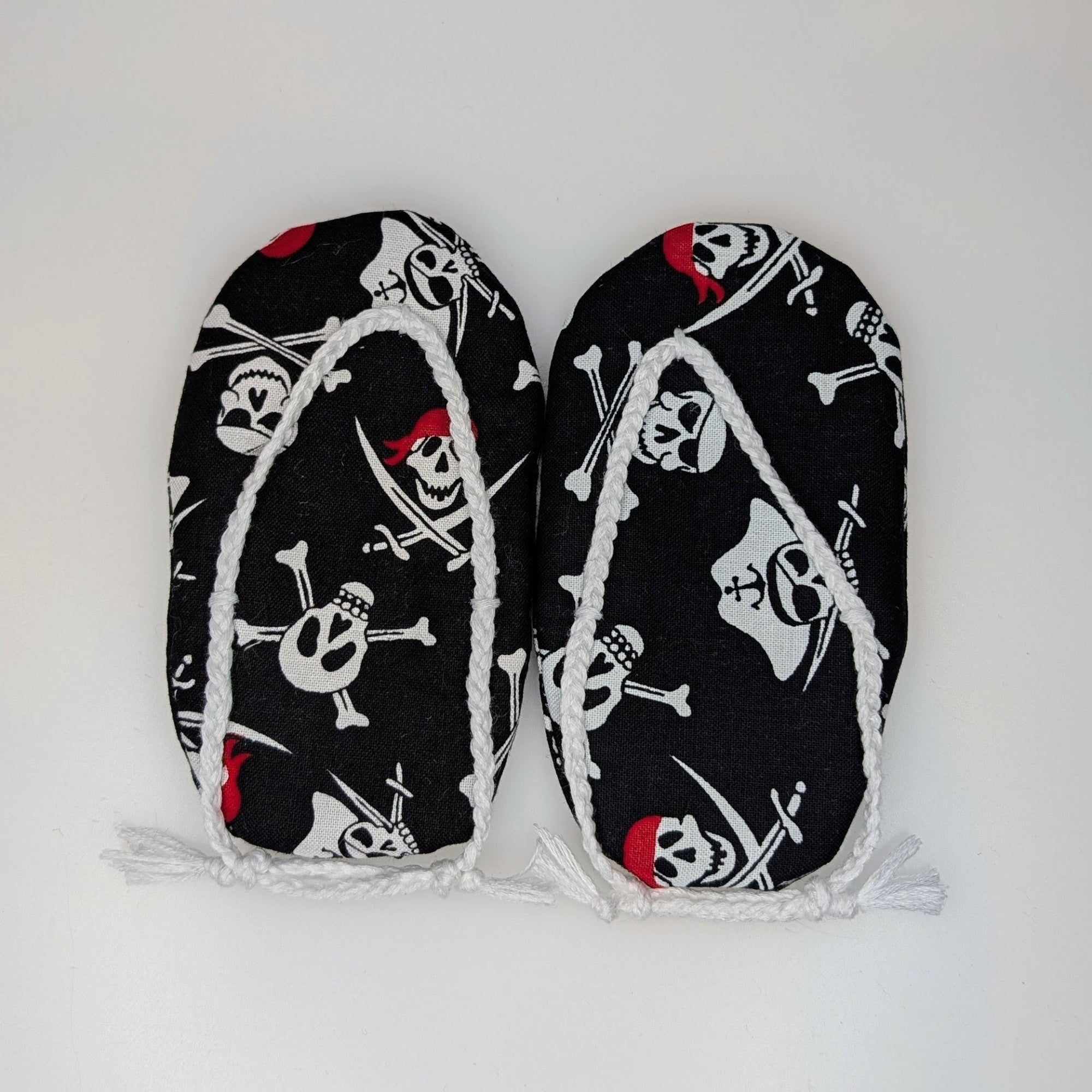 A pair of black flip-flop slippers with a white skull and crossbones pirate print and white fringe trim, laid flat on a white background.