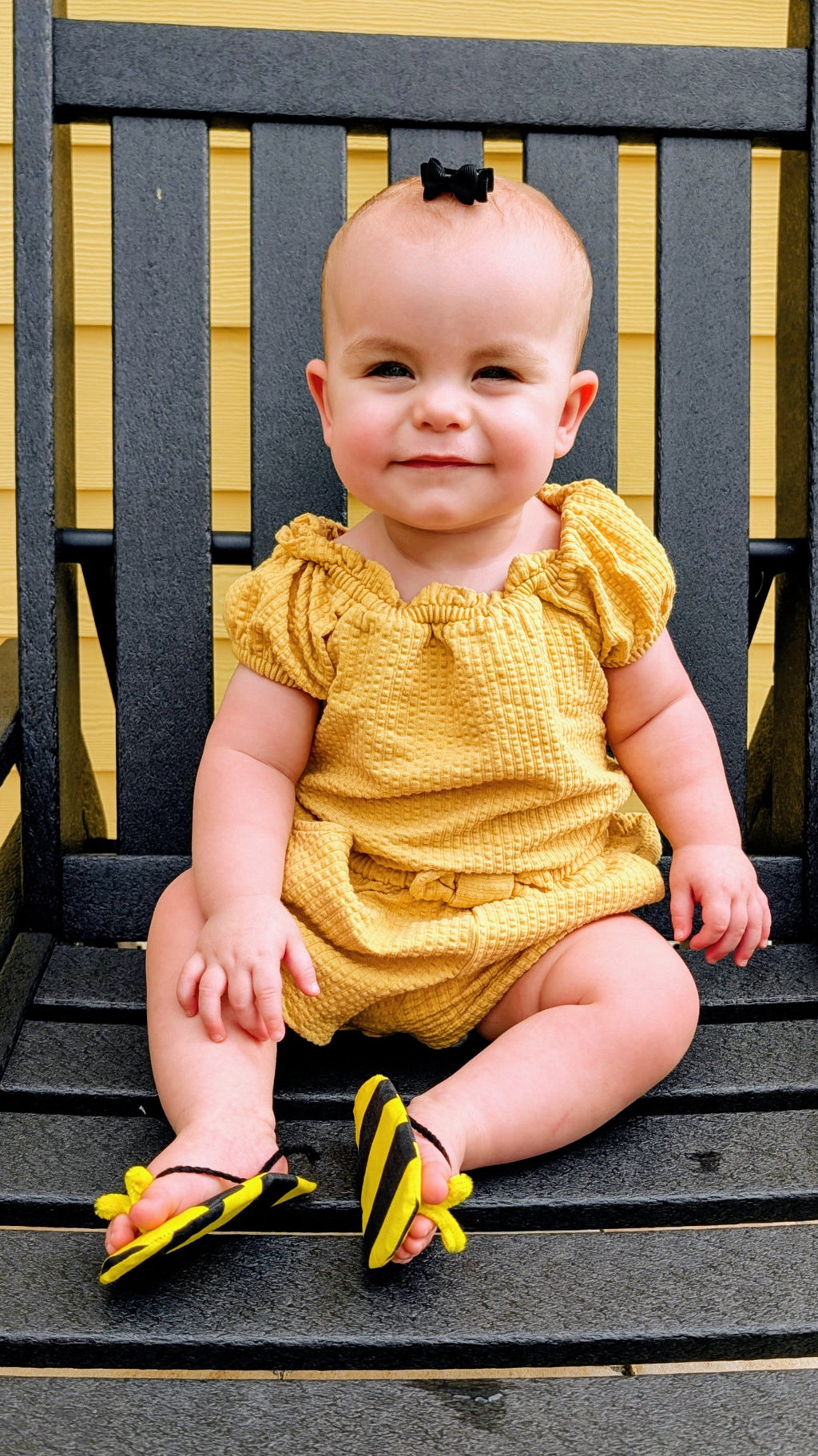 A baby girl sitting on a dark wooden bench, wearing a textured mustard yellow romper and matching yellow and black bumble-ear flip-flops.