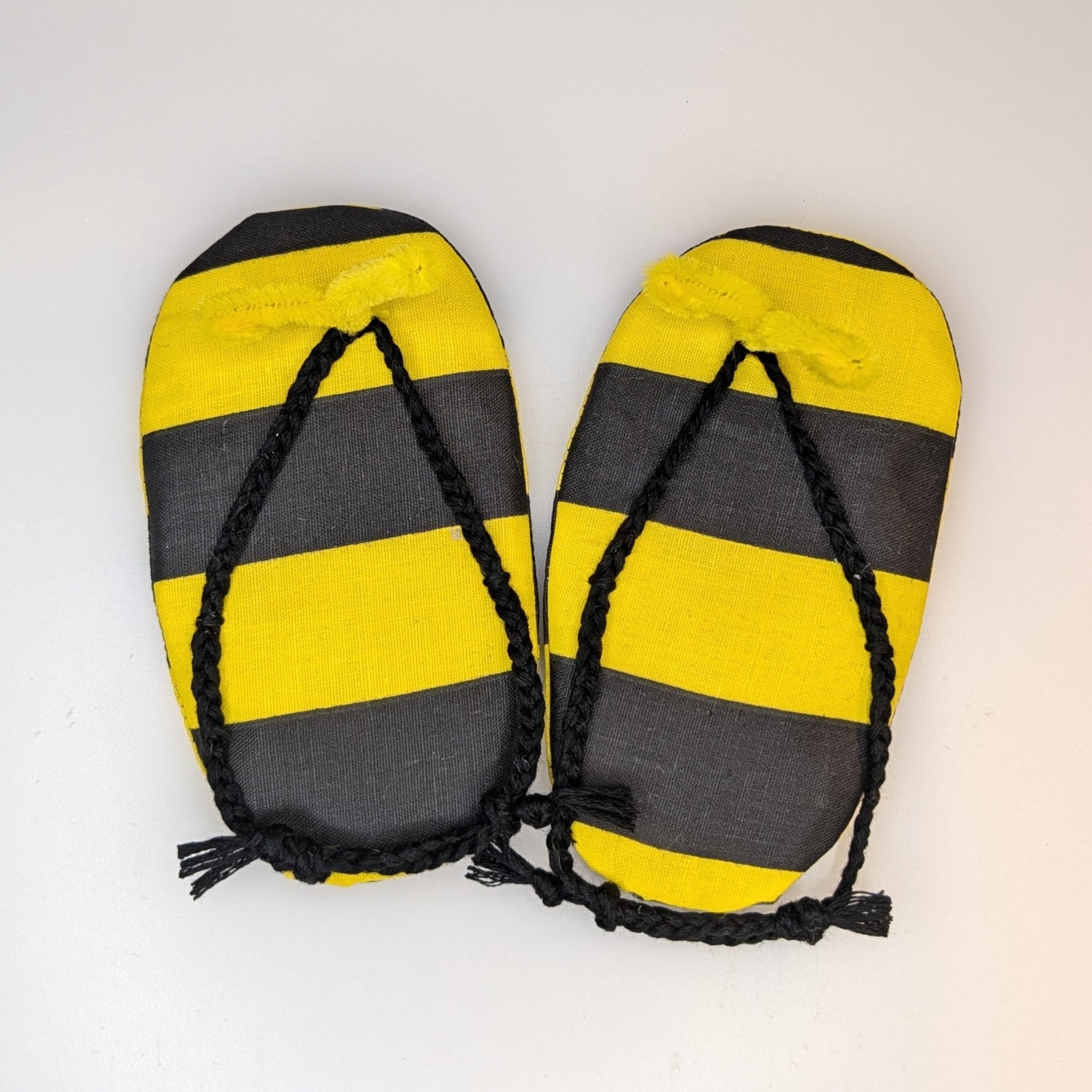 A pair of yellow and black striped flip-flops with black braided straps, laid flat on a light gray background.