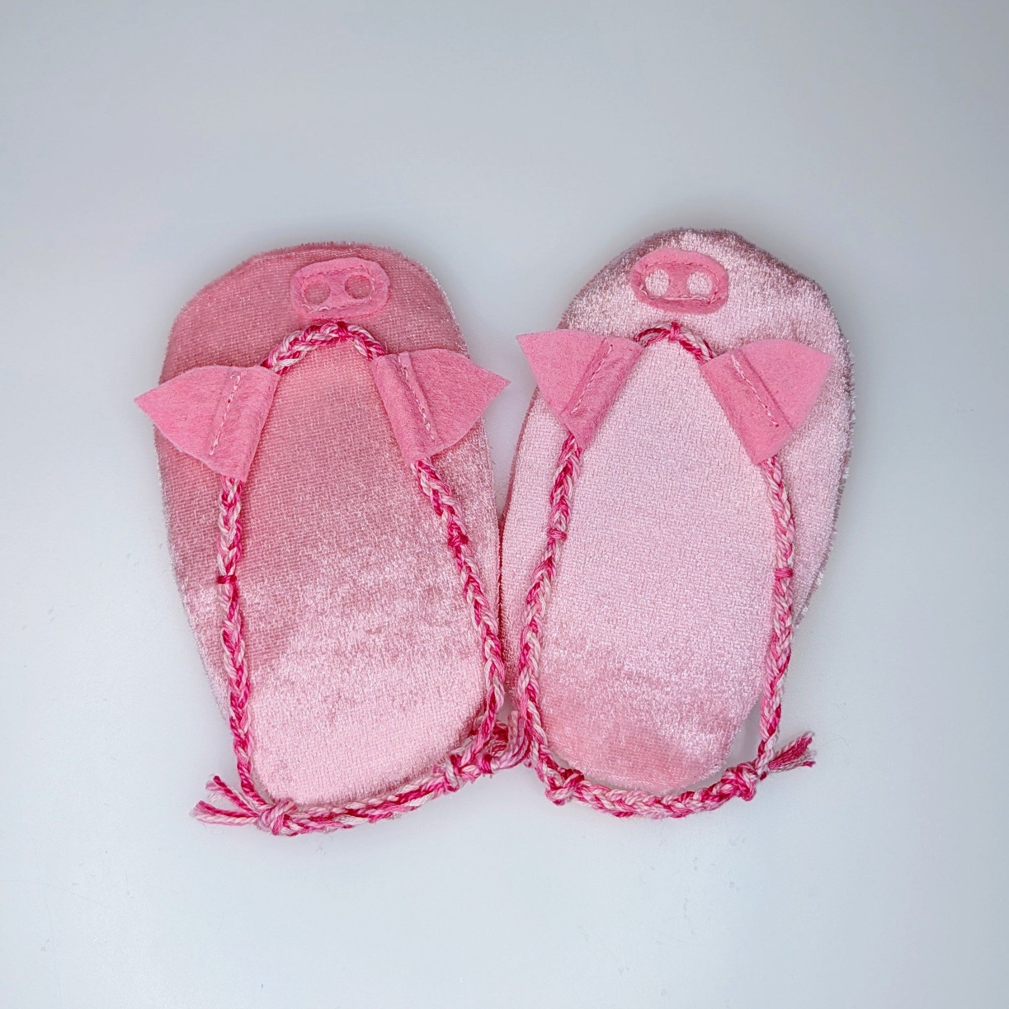 A pair of pink piggie flops with braided trim and bow details, laid flat on a light gray background.