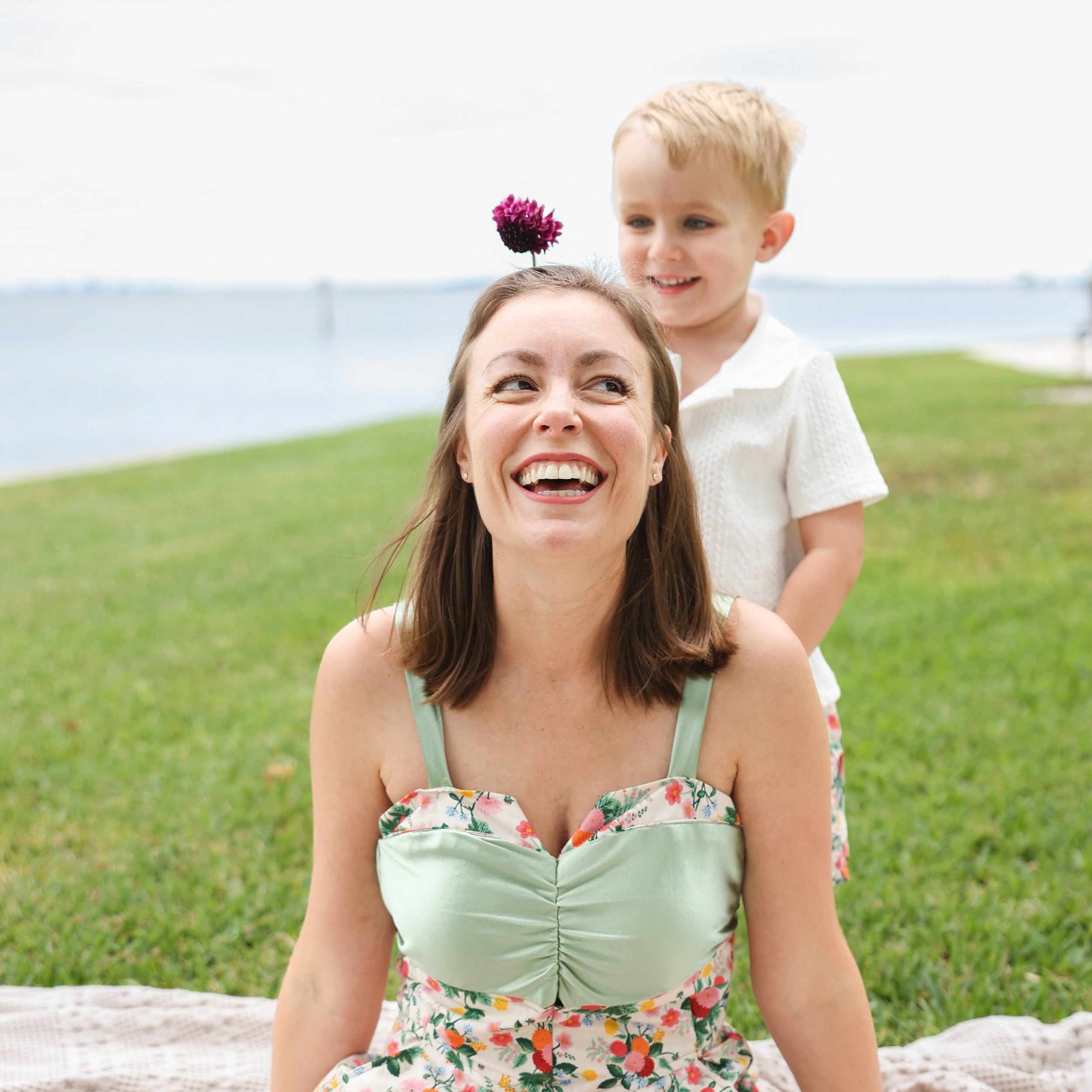 A woman and a young boy smiling outdoors on a grassy lawn near the water. The woman wears a mint green and floral sweetheart-neck dress, and the boy wears a white polo shirt.