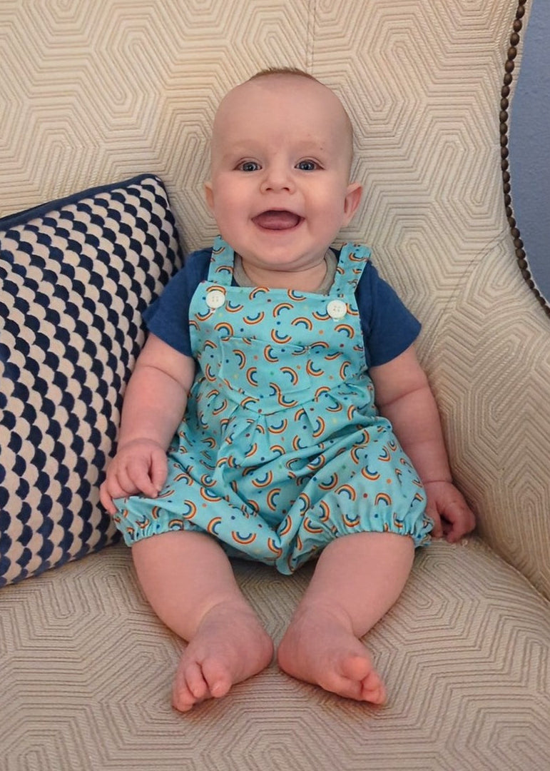 A baby sitting on a patterned beige sofa wearing a light blue raincloud print romper over a navy blue t-shirt, next to a navy and beige checkered pillow.