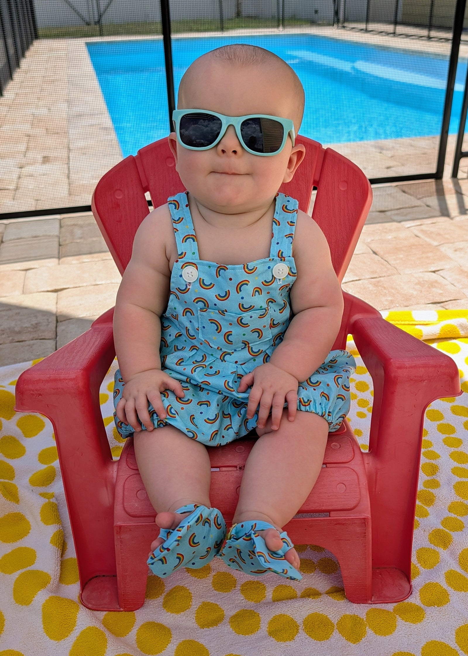A baby wearing a light blue patterned romper and matching booties sits in a red plastic chair outdoors by a swimming pool, wearing light blue-tinted sunglasses.