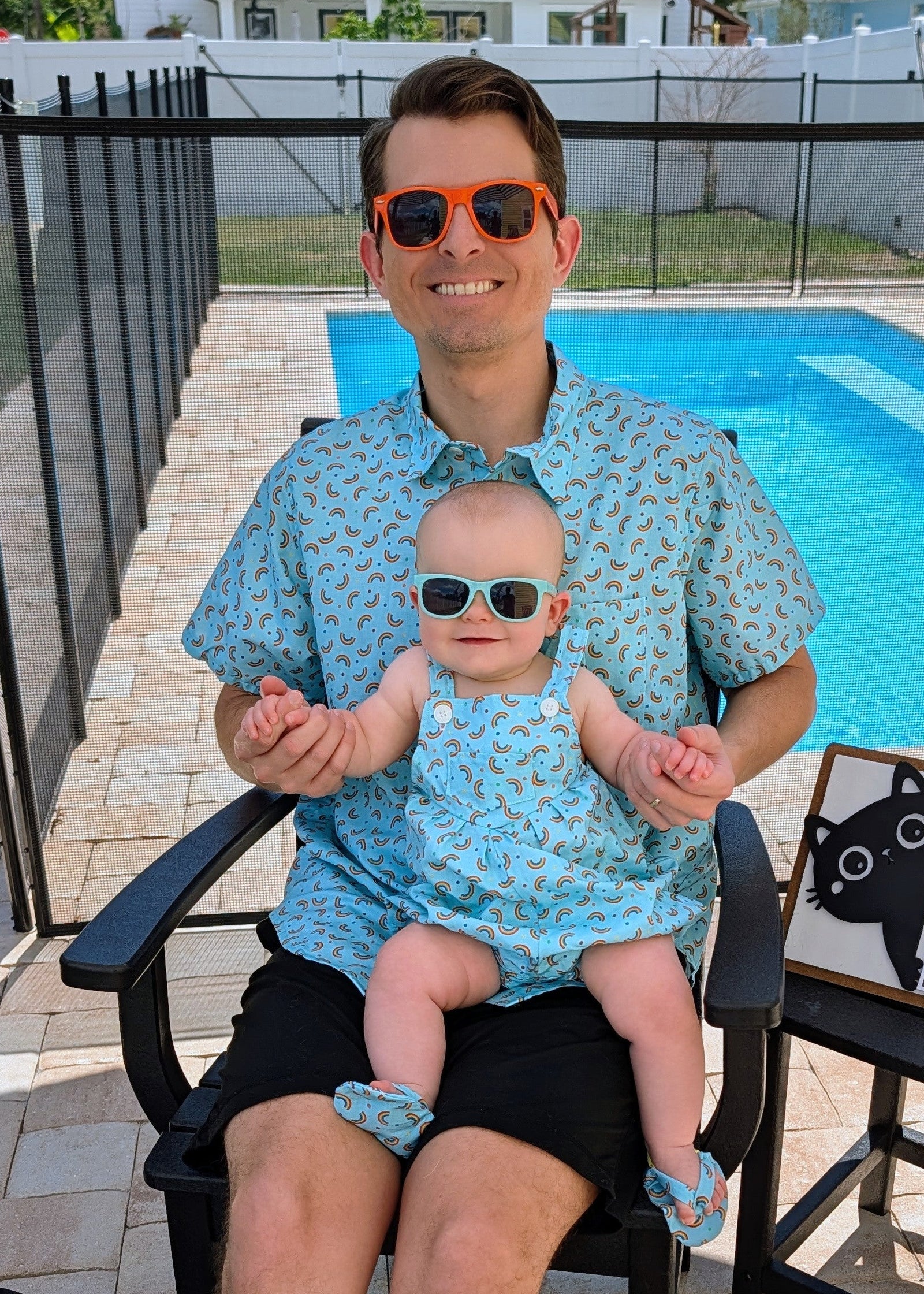 A man wearing a light blue patterned short-sleeve shirt and black shorts sits in a black outdoor chair next to a swimming pool, holding a baby dressed in a matching light blue patterned romper and matching socks. The man is wearing orange-tinted sunglasses, and a small sign on a side table reads 'Puppie 8'.