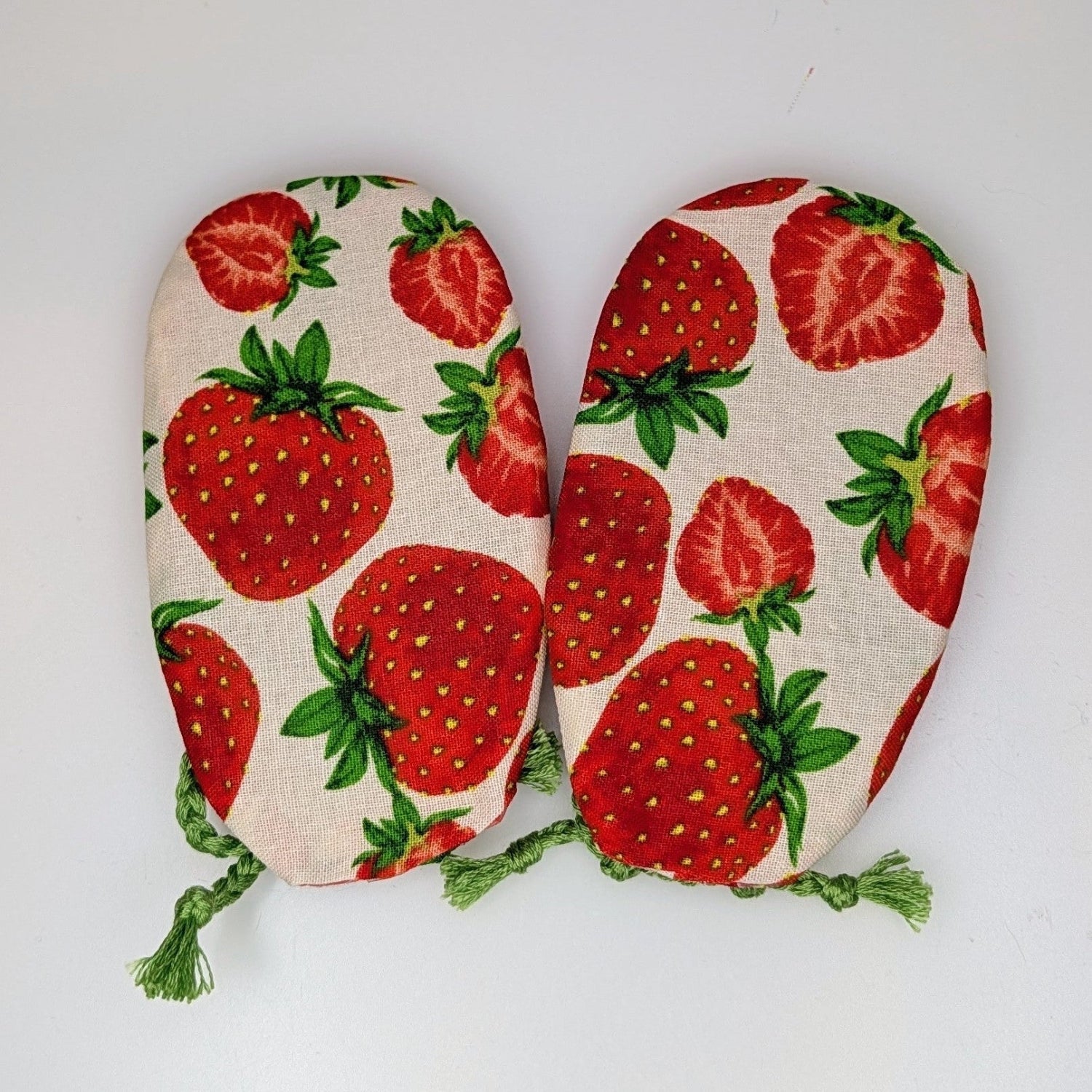 A pair of fabric oven mitts featuring a red strawberry and green leaf pattern on a cream background, with green tassels at the corners, shown on a plain white background.
