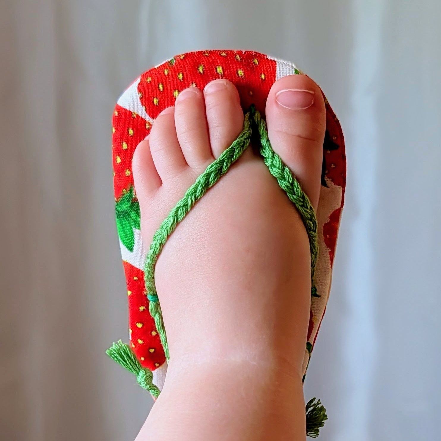 A close-up of a foot wearing a red and white strawberry-patterned thong sandal with green braided straps, set against a soft white curtain background.