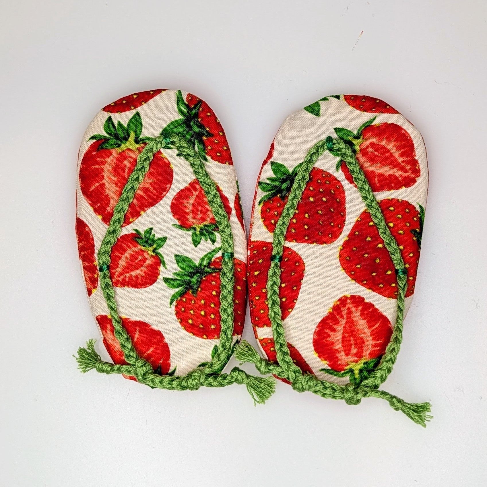 A pair of handmade fabric flip-flops featuring a red strawberry print on a cream background, with green braided straps, laid flat on a white surface.